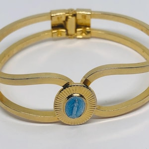 May include: A gold-tone bangle bracelet with a blue enamel oval charm featuring a depiction of the Virgin Mary. The bracelet has a simple, elegant design.