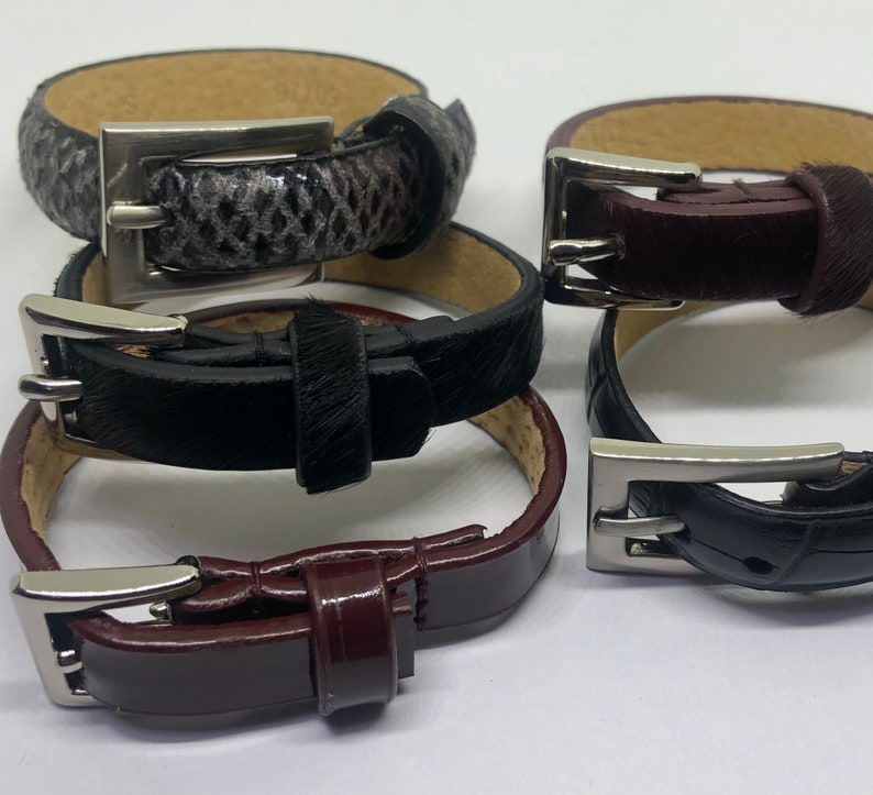 May include: Four leather bracelets with silver buckles. The bracelets are stacked on top of each other. The top bracelet is black with a brown leather strap. The second bracelet is brown with a black leather strap. The third bracelet is black with a white leather strap. The bottom bracelet is brown with a black leather strap.