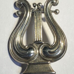 May include: A silver-toned lyre brooch with intricate details. The lyre is a musical instrument with a curved frame and strings.