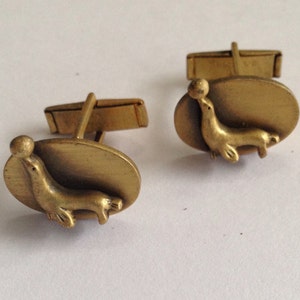 May include: A pair of gold cufflinks featuring a seal balancing a ball on its nose. The cufflinks are oval shaped and have a textured surface.