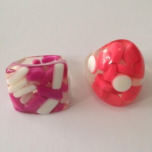 Puede incluir: Dos anillos de resina. Uno es cuadrado con formas rectangulares rosas y blancas. El otro es ovalado con formas rosas y círculos blancos. Ambos anillos tienen una base transparente.