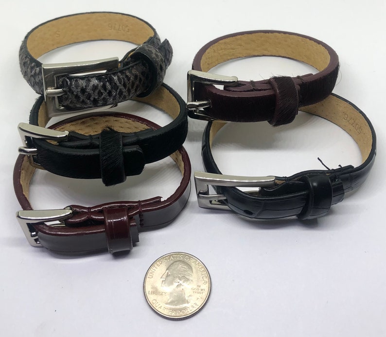 May include: Four leather bracelets with silver buckles. The bracelets are stacked on top of each other. The bracelets are in different colors, including black, brown, and gray.