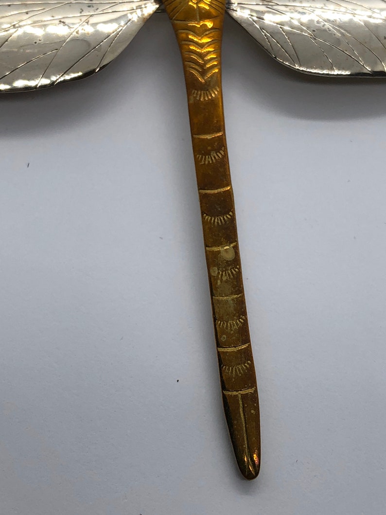 Puede incluir: Un broche de lib&eacute;lula de oro con alas de plata. El cuerpo de la lib&eacute;lula est&aacute; decorado con intrincados patrones.