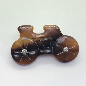 May include: A vintage-style brooch in the shape of a bicycle. The brooch is primarily brown with a marbled effect, featuring two wheels with crystal accents and silver-colored studs. The design is reminiscent of antique jewelry.