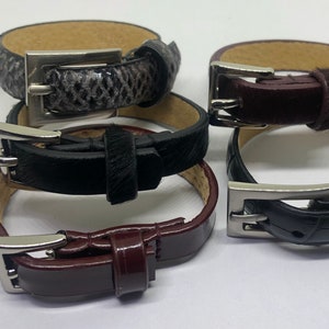 May include: Four leather bracelets with silver buckles. The bracelets are stacked on top of each other. The top bracelet is black with a brown leather strap. The second bracelet is brown with a black leather strap. The third bracelet is black with a white leather strap. The bottom bracelet is brown with a black leather strap.