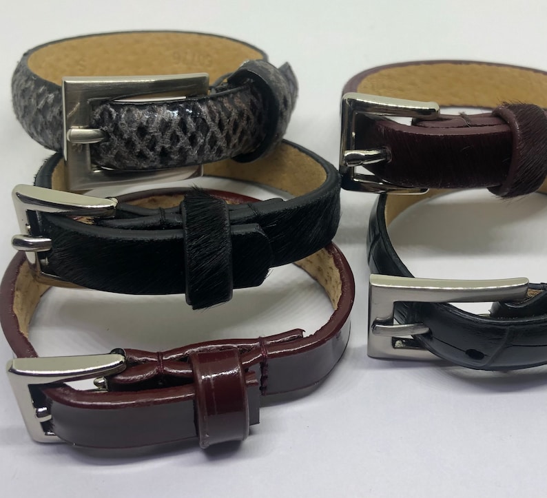 May include: Four leather bracelets with silver buckles. The bracelets are stacked on top of each other. The top bracelet is black with a brown leather strap. The second bracelet is brown with a black leather strap. The third bracelet is black with a white leather strap. The bottom bracelet is brown with a black leather strap.