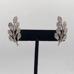 May include: A pair of silver-toned leaf-shaped earrings. Each earring features a stem with multiple textured leaves. The earrings are displayed on a black stand against a white background. The earrings are a classic jewelry item.