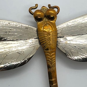 Puede incluir: Un broche de lib&eacute;lula de plata y oro con alas detalladas y un cuerpo texturizado.