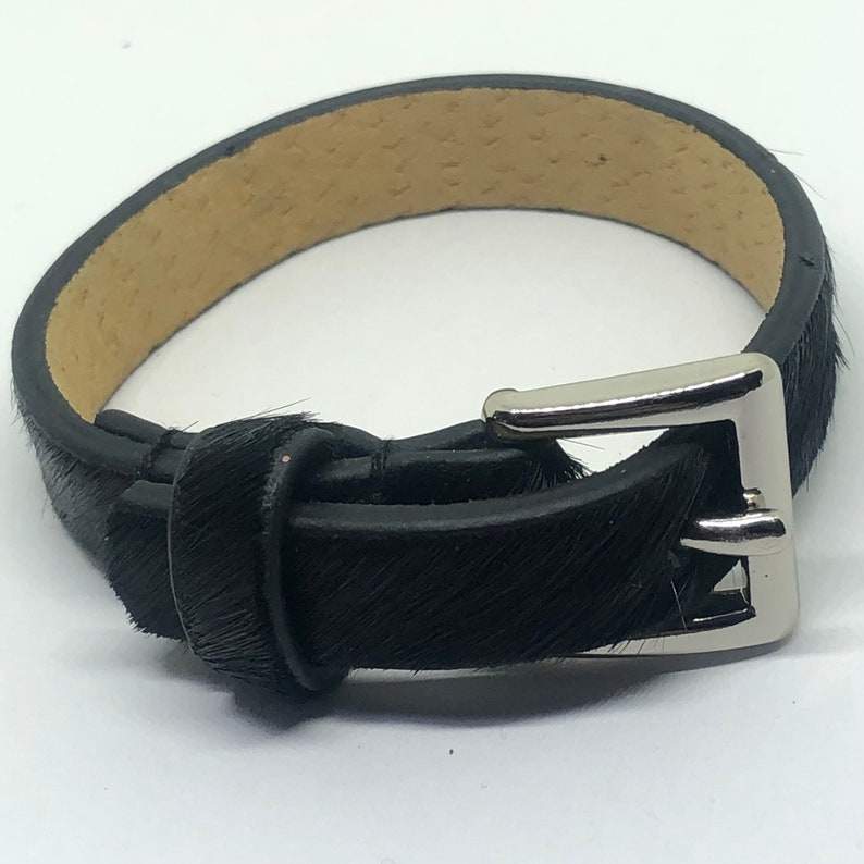 May include: A black leather bracelet with a silver buckle. The bracelet is made of hair-on hide leather.