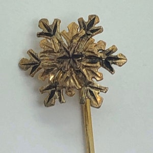 May include: A gold snowflake pin with a detailed design. The pin is attached to a thin gold stem.