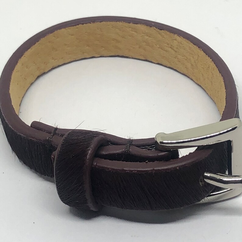 May include: A brown leather bracelet with a silver buckle. The bracelet is made of a soft, hairy leather.