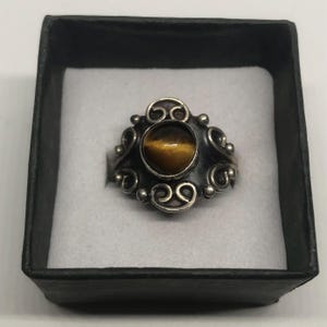 May include: A silver-toned ring with an oval, tiger's eye stone set in the center. The ring has an ornate design with swirling patterns and small, round accents. The ring is displayed in a black box with a white interior.
