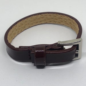 May include: A dark brown leather bracelet with a silver buckle. The bracelet is made of a smooth, polished leather and has a simple, elegant design.