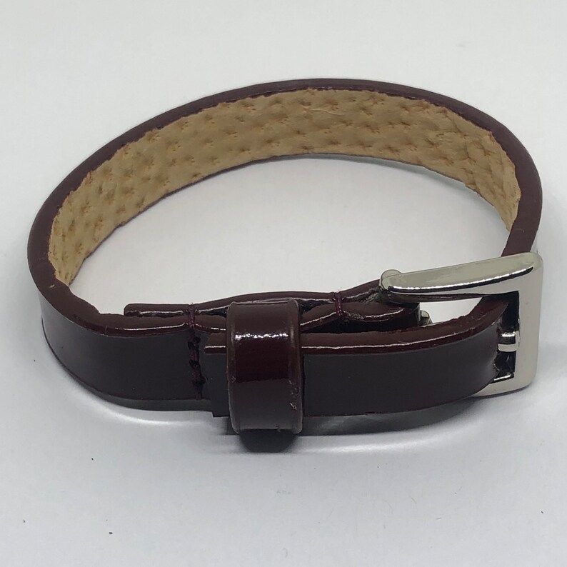 May include: A dark brown leather bracelet with a silver buckle. The bracelet is made of a smooth, polished leather and has a simple, elegant design.