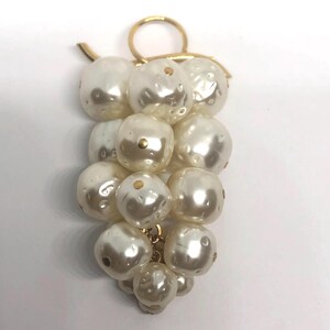 May include: A cluster of faux pearl beads in the shape of grapes, with a gold-tone metal stem and ring. The off-white pearls have a slightly irregular shape and small gold accents. A unique jewelry piece.