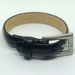May include: A black leather watch band with a silver buckle. The band has a crocodile pattern and the buckle has a simple design.