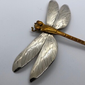 Puede incluir: Un broche de lib&eacute;lula de plata con detalles dorados. Las alas est&aacute;n detalladas con l&iacute;neas intrincadas y el cuerpo es de color dorado s&oacute;lido.