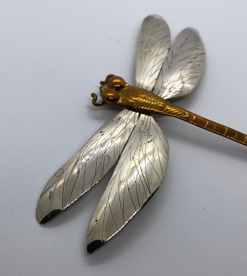 Puede incluir: Un broche de lib&eacute;lula de plata con detalles dorados. Las alas est&aacute;n detalladas con l&iacute;neas intrincadas y el cuerpo es de color dorado s&oacute;lido.