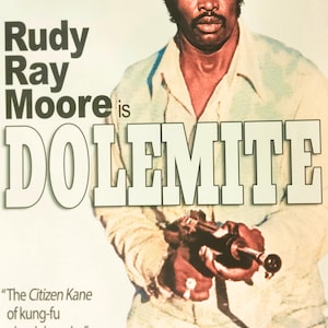 May include: Poster for the film "Dolemite" featuring Rudy Ray Moore. The image shows Moore wearing a light-colored shirt and hat, holding a prop gun. Text includes "The Citizen Kane of kung-fu pimpin' movies" and "The New York Times."