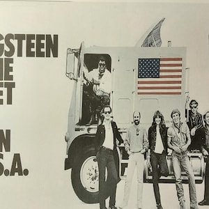 May include: A black and white poster advertising a Bruce Springsteen and the E Street Band concert tour. The poster features a large semi-truck with an American flag on the side and the band members standing in front of it. The text on the poster reads "Bruce Springsteen and the E Street Band Born in the U.S.A. Tour."