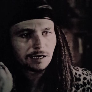 May include: A man with long, dark hair and a black bandana is giving the middle finger. He is wearing a leopard print shirt.