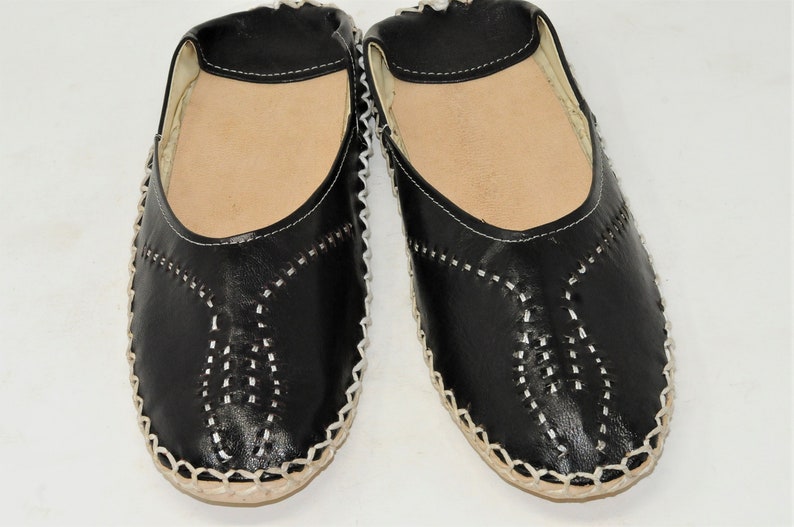 Pode incluir: Um par de sapatos pretos de couro sem cadar&ccedil;os com costuras brancas e uma palmilha castanha clara. Os sapatos t&ecirc;m bico arredondado e um padr&atilde;o costurado decorativo na g&aacute;spea.