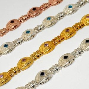 May include: Close-up of four ornate bracelets in rose gold, silver, and gold tones. Each bracelet features oval-shaped links with filigree designs and a central gemstone in various colors, including black, blue, and pink.