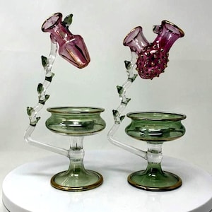 Könnte beinhalten: Zwei dekorative Kerzenhalter aus Glas. Jeder hat einen grünen Sockel mit Goldrand, einen klaren Stiel und einen astähnlichen Arm, der eine rosa vasenförmige Spitze hält. Eine Spitze ist glatt, die andere strukturiert.