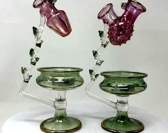 May include: Two glass candle holders with green bases and stems featuring small leaves. Each holder has a clear glass bowl for a tea light and an attached curved arm supporting a pink glass flower-shaped top, one smooth and one with gold dots.