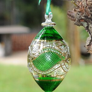 May include: A green glass Christmas bauble with gold and white detailing. The bauble is egg-shaped and has a loop at the top for hanging.