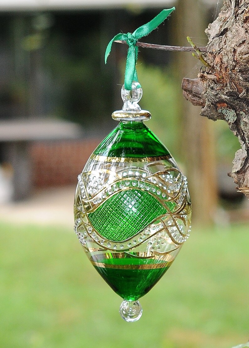 May include: A green glass Christmas bauble with gold and white detailing. The bauble is egg-shaped and has a loop at the top for hanging.
