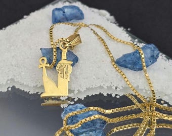 May include: A gold-colored necklace featuring a pendant shaped like an Egyptian pharaoh in profile holding an ankh symbol. The pendant is attached to a delicate box chain, showcasing a polished metallic finish and intricate engraved details.