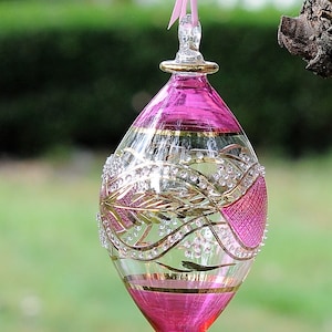 May include: A pink glass bauble with gold and white detailing. The bauble is hanging from a pink ribbon.