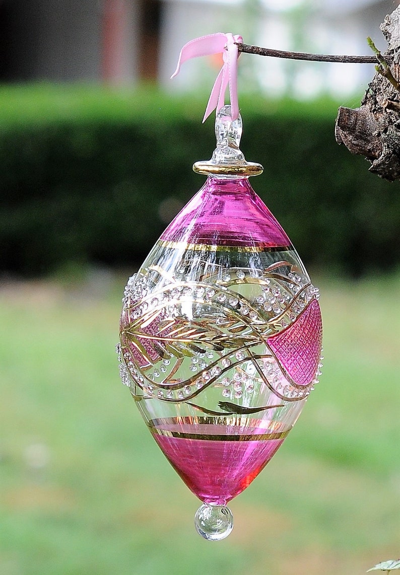 May include: A pink glass bauble with gold and white detailing. The bauble is hanging from a pink ribbon.