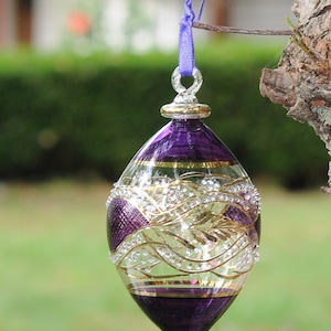 May include: A purple glass bauble with gold and white detailing. The bauble is egg-shaped and has a loop at the top for hanging.