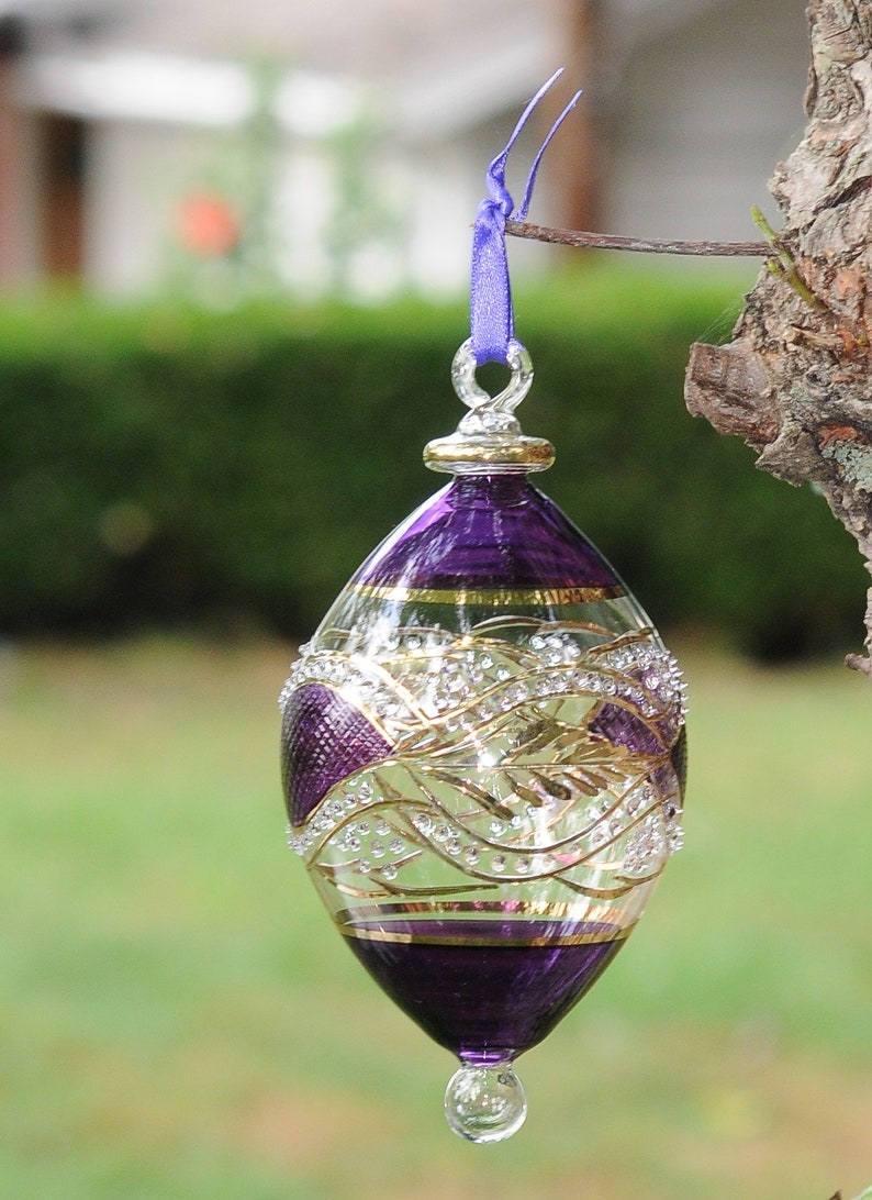 May include: A purple glass bauble with gold and white detailing. The bauble is egg-shaped and has a loop at the top for hanging.