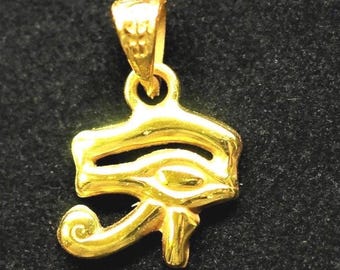 May include: A gold-colored pendant in the shape of the Eye of Horus, a symbol of protection and royal power in ancient Egypt. The pendant has a loop at the top for a chain and is set against a dark background.