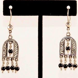 May include: A pair of silver-toned dangle earrings. Each earring features an arched filigree design with a central black bead and a cluster of three dangling black beads. The earrings are displayed against a black stand.