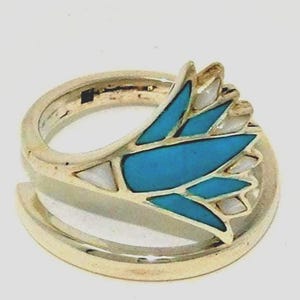 May include: A gold-colored ring with a turquoise and white inlay design. The ring features a stylized floral or feather motif with bright blue and white segments. The ring is set against a plain white background.