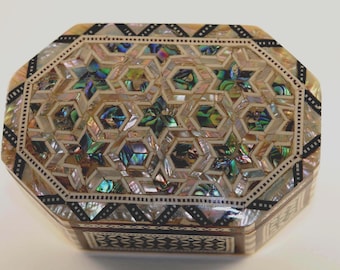 May include: An octagonal decorative box with a mother-of-pearl inlay design. The box features a geometric pattern of stars and hexagons, with iridescent accents. The edges are trimmed with black and white detailing, creating a striking contrast.