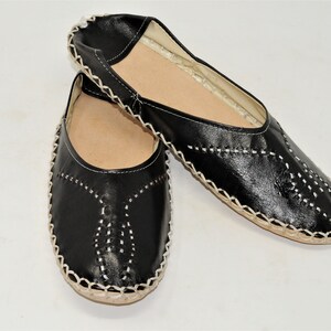 Pode incluir: Um par de sapatos pretos de couro sem cadar&ccedil;os com detalhes de costura brancos. Os sapatos t&ecirc;m bico redondo e sola de cor natural. As costuras formam padr&otilde;es decorativos na g&aacute;spea.