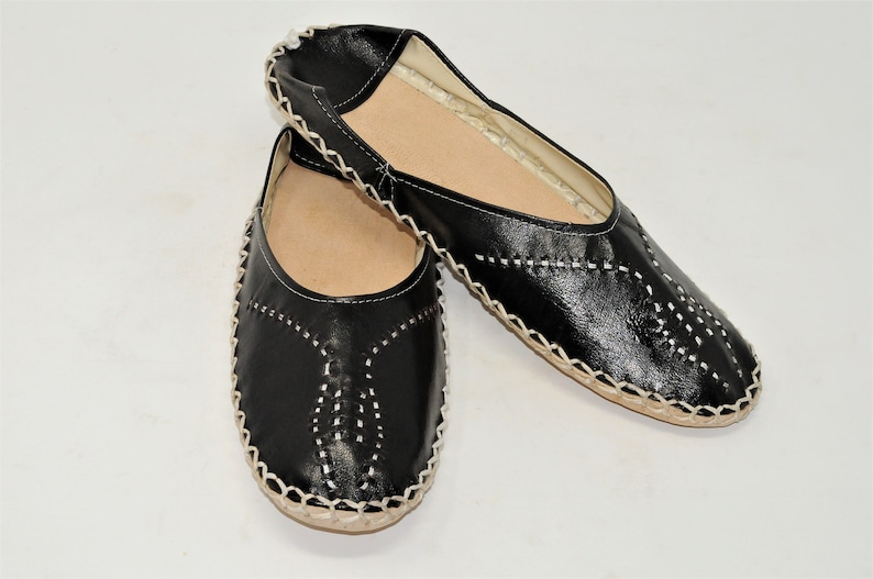 Pode incluir: Um par de sapatos pretos de couro sem cadar&ccedil;os com detalhes de costura brancos. Os sapatos t&ecirc;m bico redondo e sola de cor natural. As costuras formam padr&otilde;es decorativos na g&aacute;spea.