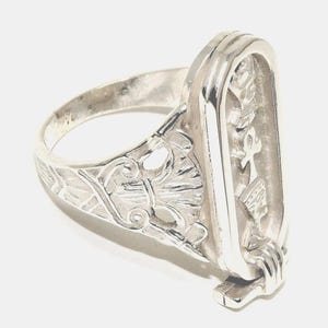 May include: A silver ring with an Egyptian cartouche design. The ring features an oval centerpiece with hieroglyphic symbols and a decorative band with floral and scrollwork details. The ring is made of a shiny, reflective metal.