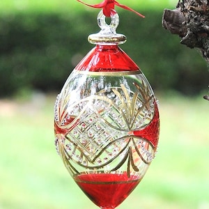 May include: An oval-shaped glass ornament featuring a clear center with gold painted geometric lines and small clear crystals. The top and bottom sections are solid red glass with gold trim. A red ribbon is tied to the top loop for hanging.
