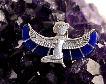 May include: A silver-toned metal brooch featuring a kneeling winged figure with outstretched wings. The wings are inlaid with deep blue segments. The figure has a profile view of a head and torso, with detailed wing feathers and a smooth finish.