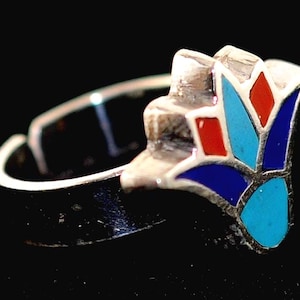 May include: Silver-tone ring with a floral design. The ring features a cluster of red, blue, and turquoise-colored geometric shapes. The ring is set against a black background.
