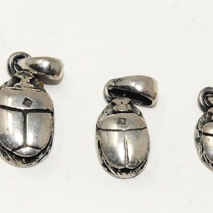 May include: Three silver-toned metal pendants shaped like scarab beetles, arranged in a row from largest to smallest. Each pendant features a smooth, rounded back with a central line and a small square detail, attached to a sturdy metal bail loop.
