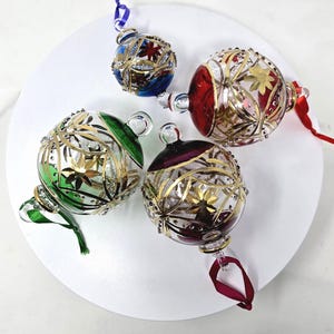 Puede incluir: Cuatro adornos esféricos de cristal con secciones transparentes, motivos florales pintados en oro y segmentos de cristal de colores azul, rojo, verde y morado. Cada adorno incluye una pequeña anilla de cristal con una cinta a juego.