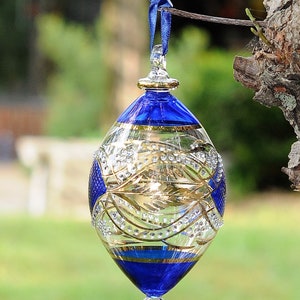 May include: A blue and gold glass bauble with a delicate design and a clear glass drop. The bauble is hanging from a blue ribbon.