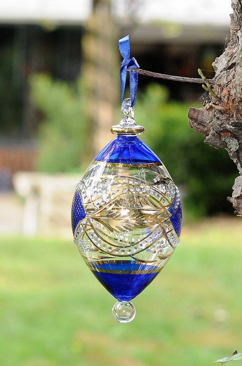 May include: A blue and gold glass bauble with a delicate design and a clear glass drop. The bauble is hanging from a blue ribbon.